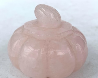 May include: A carved rose quartz pumpkin figurine. The light pink gemstone is sculpted with a ribbed body and a curved stem. The pumpkin is a decorative item, likely for display or as a collectible.