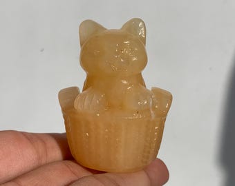 May include: A carved, light orange cat figurine sitting in a basket. The cat has pointed ears and a smiling face. The basket has a textured, woven appearance. The figurine is made of a translucent material.