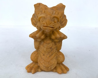 May include: A small, tan-colored stone sculpture of a mythical creature. The figure has a detailed face with large eyes and a small mouth. It has large ears and is posed with its arms at its sides. The sculpture is approximately 3 inches tall.