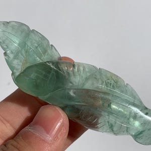 May include: A carved, translucent green fluorite leaf sculpture. The leaf has detailed veining and a smooth, polished finish. The sculpture is held in a hand against a white background.