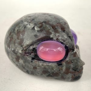 May include: A grey stone alien head sculpture with pink and purple eye-shaped gemstones. The sculpture has a smooth, polished finish and a detailed profile. The alien head is a unique decorative item.
