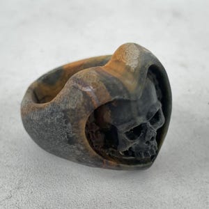 May include: A dark, carved ring featuring a skull design. The ring has a hooded shape with a detailed skull face. The ring's colour is a mix of dark grey, brown, and black.