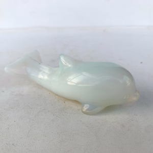 May include: A pearlescent white dolphin ornament. The dolphin has a smooth, rounded body with a dorsal fin and a tail fin. The ornament is made of a translucent material, giving it a subtle glow. The dolphin is lying on its side.