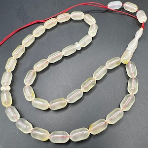 Puede incluir: Un collar de cuentas de oración de color claro con cuentas oblongas y un cordón rojo. Las cuentas son translúcidas con un sutil tono amarillo, y el cordón es de un rojo vibrante. El collar está sobre una superficie oscura.