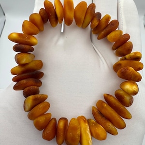 Puede incluir: Una pulsera hecha de piedras de ámbar pulidas. Las piedras de ámbar son de varias formas y tamaños, con tonos que van del naranja claro al oscuro y marrón. La pulsera está ensartada, creando una forma circular.