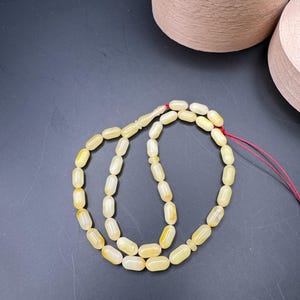 May include: A strand of light yellow prayer beads with an oval shape. The beads are strung on a red string. The background is a dark gray surface with two light brown containers.