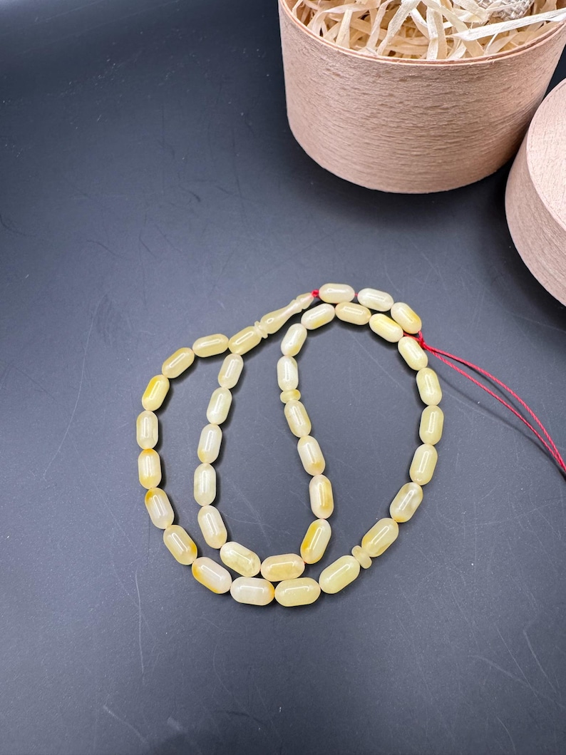 May include: A strand of light yellow prayer beads with an oval shape. The beads are strung on a red string. The background is a dark gray surface with two light brown containers.
