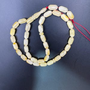 May include: A strand of prayer beads with oval-shaped, off-white beads with yellow accents. The beads are strung on a red cord and have a decorative, elongated bead at the top. The background is a dark, solid color.