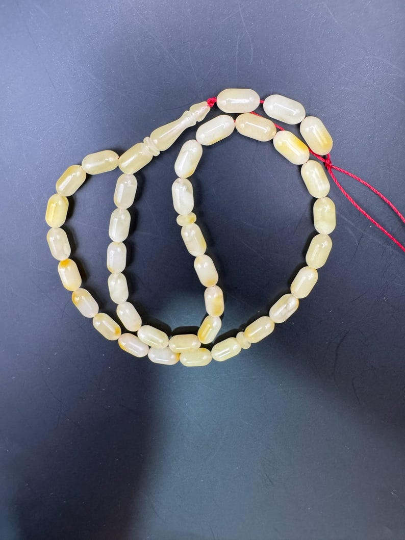 May include: A strand of prayer beads with oval-shaped, off-white beads with yellow accents. The beads are strung on a red cord and have a decorative, elongated bead at the top. The background is a dark, solid color.