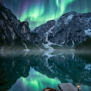 Può includere: Una barca a remi in legno chiamata AURORA è ormeggiata a un molo di legno su un lago calmo. La scena presenta imponenti montagne spolverate di neve che si riflettono nell'acqua sotto un cielo notturno vibrante con aurore boreali verdi e viola.