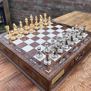 May include: A wooden chess set with gold and silver chess pieces arranged on a checkered board. The board is inlaid with light-colored squares and framed by a decorative border. The chess pieces are in place, ready for a game.