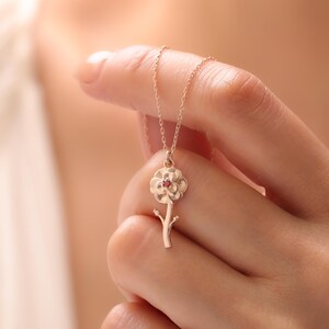 May include: A gold-colored flower pendant necklace with a small red gemstone in the center. The flower is on a stem with two small leaves. The necklace is held by a person.