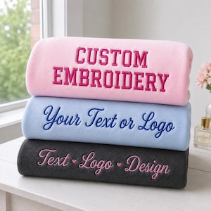 May include: A stack of three folded fleece blankets. The top blanket is light pink with the dark pink embroidered text CUSTOM EMBROIDERY. The middle blanket is light blue with dark blue embroidered text Your Text or Logo. The bottom blanket is dark gray with light pink embroidered text Text • Logo • Design.