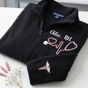 May include: Black fleece zip-up jacket with a pink stethoscope design and the name "Chloe, RN" embroidered on the front. A pink caduceus symbol is embroidered on the sleeve. The jacket is folded on a white surface.