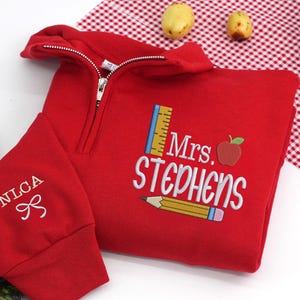 May include: A red quarter-zip sweatshirt with the name "Mrs. Stephens" embroidered in white, along with a ruler, apple, and pencil design. The left sleeve has "NLCA" and a bow embroidered in white.
