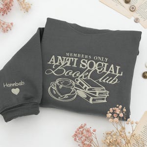 Custom embroidered "Anti Social Book Club" Gildan Sweatshirt in Dark Heather. Features a coffee cup and books design thimbled in cream thread. Personalized "Hannbab" name and heart on the sleeve. Styled with pink dried flowers, a gold locket, and vintage book pages. Perfect bookish gift for introverts, readers, and library lovers seeking aesthetic oversized sweaters.