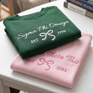 May include: A dark green sweatshirt with white embroidered text reading Sigma Phi Omega EST 1998 and a bow icon, stacked on a light pink sweatshirt with pink embroidered text reading Alpha Theta Tau EST 2004 and a bow icon.