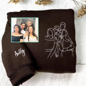 Puede incluir: Una sudadera marrón oscuro con una fotografía enmarcada de cuatro personas y un contorno bordado en blanco de un grupo de personas. El nombre "Aully" está bordado en blanco en la manga.
