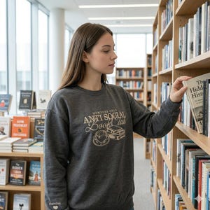 A young woman browsing library shelves while wearing a custom "Anti Social Book Club" sweatshirt in Dark Heather. The camera captures the personalized sleeve embroidery as she reaches for a book. Professional library-themed photography. Perfect for SEO targeting bookish lifestyle, librarian style, and personalized reading-themed clothing on Etsy.