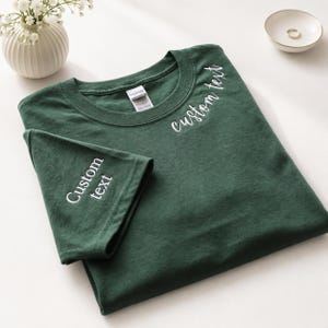 May include: A dark forest green short-sleeve t-shirt featuring white embroidered text that reads "custom text" on the upper left chest area and on the sleeve. The shirt has a crew neckline and a Gildan brand label visible at the inner collar.