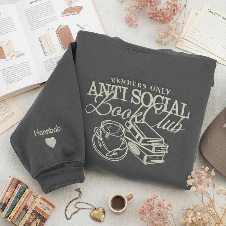 Aesthetic flat lay of a charcoal "Anti Social Book Club" sweatshirt surrounded by miniature books, a tiny coffee cup, a gold necklace, and dried flowers. The composition features the personalized "Hannbab" sleeve prominently. High-resolution 8K photography for Etsy. Best for "Bookworm Gift Idea," "Customized Reading Sweatshirt," and "Aesthetic Reader Apparel."