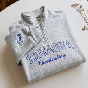 May include: A gray sweatshirt with a quarter-zip neckline. The word "TAMAQUA" is embroidered in blue block letters, with "Cheerleading" below. The left sleeve cuff has the word "Coach" embroidered in blue script.