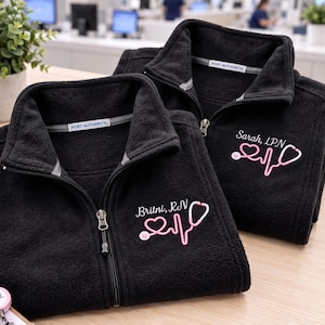 May include: Two black fleece zip-up jackets featuring the Port Authority brand label. Each jacket is embroidered with a pink stethoscope forming a heart and heartbeat line, with the names Britni, RN and Sarah, LPN stitched above the design.