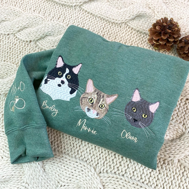 Heather Dark Green embroidered sweatshirt with three custom cat faces and names—Bradley, Minnie, Owen—plus infinity symbol on sleeve. Personalized cat mom sweatshirt, custom pet lover gift with embroidery.