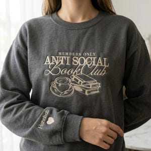 A woman wearing a custom Dark Heather "Anti Social Book Club" embroidered sweatshirt. The cream-colored bookish design is prominently displayed on the chest. Realistic product mockup for American lifestyle aesthetic. Great for marketing oversized comfort wear, reader-themed apparel, and cozy gifts for book club members or English teachers.