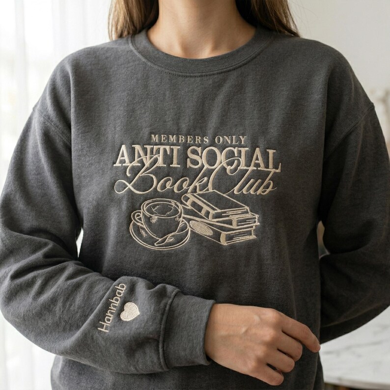 A woman wearing a custom Dark Heather "Anti Social Book Club" embroidered sweatshirt. The cream-colored bookish design is prominently displayed on the chest. Realistic product mockup for American lifestyle aesthetic. Great for marketing oversized comfort wear, reader-themed apparel, and cozy gifts for book club members or English teachers.