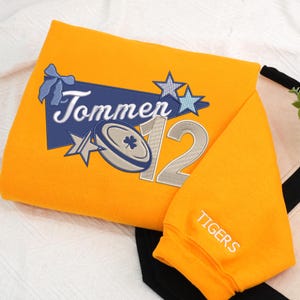 May include: Mustard yellow sweatshirt with embroidered design. Features the name "Tommen" above a rugby ball and the number "12" in silver. The sleeve reads "TIGERS" in white. A blue bow and stars complete the design.