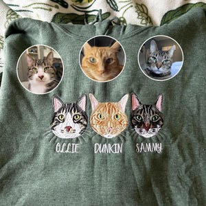 Personalized embroidered heather dark green sweatshirt with three custom cat faces—tabby cat, orange cat, and dark striped cat—plus names Ollie, Dunkin, and Sammy. Custom pet portrait embroidery gift for cat moms and dads.