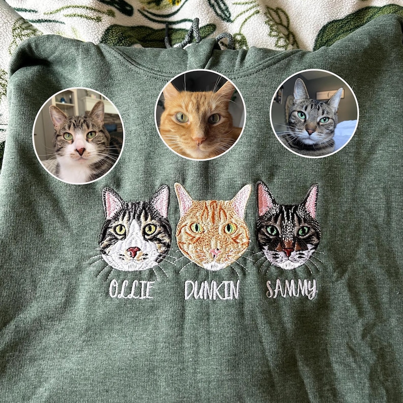 Personalized embroidered heather dark green sweatshirt with three custom cat faces—tabby cat, orange cat, and dark striped cat—plus names Ollie, Dunkin, and Sammy. Custom pet portrait embroidery gift for cat moms and dads.