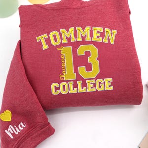 May include: A maroon hoodie with the text "TOMMEN 13 COLLEGE" in yellow. The name "Kavanagh" is vertically aligned on the left side of the number 13. The sleeve has the name "Mia" and a yellow heart.