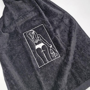 May include: Dark gray towel with a white outline illustration of a person in a swimsuit holding a towel. The illustration is in a white rectangular frame. The towel is folded and has a soft, plush texture.