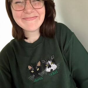 Personalized embroidered cat sweatshirt in forest green, worn by smiling woman. Features two custom cat portraits with names Sheva and Sybil. Custom pet lover sweater, unique embroidered cat mom gift.