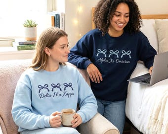 May include: Two women wearing light blue and navy blue sweatshirts. The light blue sweatshirt has the text "Delta Mu Kappa" and three bow graphics. The navy blue sweatshirt has the text "Alpha Xi Omicron" and three bow graphics. Both sweatshirts are long-sleeved.