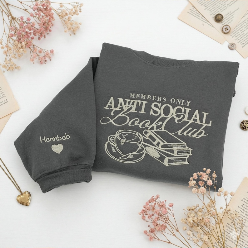 Custom embroidered "Anti Social Book Club" Gildan Sweatshirt in Dark Heather. Features a coffee cup and books design thimbled in cream thread. Personalized "Hannbab" name and heart on the sleeve. Styled with pink dried flowers, a gold locket, and vintage book pages. Perfect bookish gift for introverts, readers, and library lovers seeking aesthetic oversized sweaters.