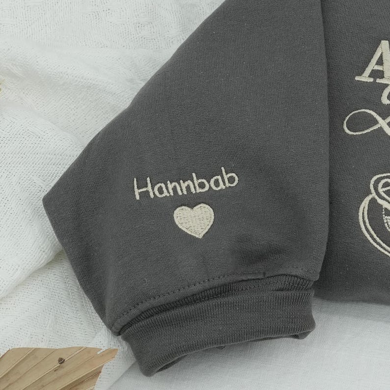 Detailed close-up of personalized sleeve embroidery on a Dark Heather sweatshirt. Features the name "Hannbab" and a small heart thimbled in cream thread. Pure fabric appearance under 6500K daylight. Perfect visual for "Custom Name Sweatshirt" and "Personalized Sleeve Embroidery" keywords, showing the fine detail and high-resolution stitching quality.