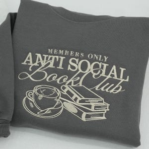 Macro detail of "Members Only Anti Social Book Club" embroidery on a charcoal grey Gildan sweatshirt. High-quality cream thread stitching showcases a realistic 3D relief effect of a coffee cup and stacked books. Professional commercial photography highlights the dense thread texture and premium craftsmanship. Ideal for showcasing custom embroidery quality on Etsy.
