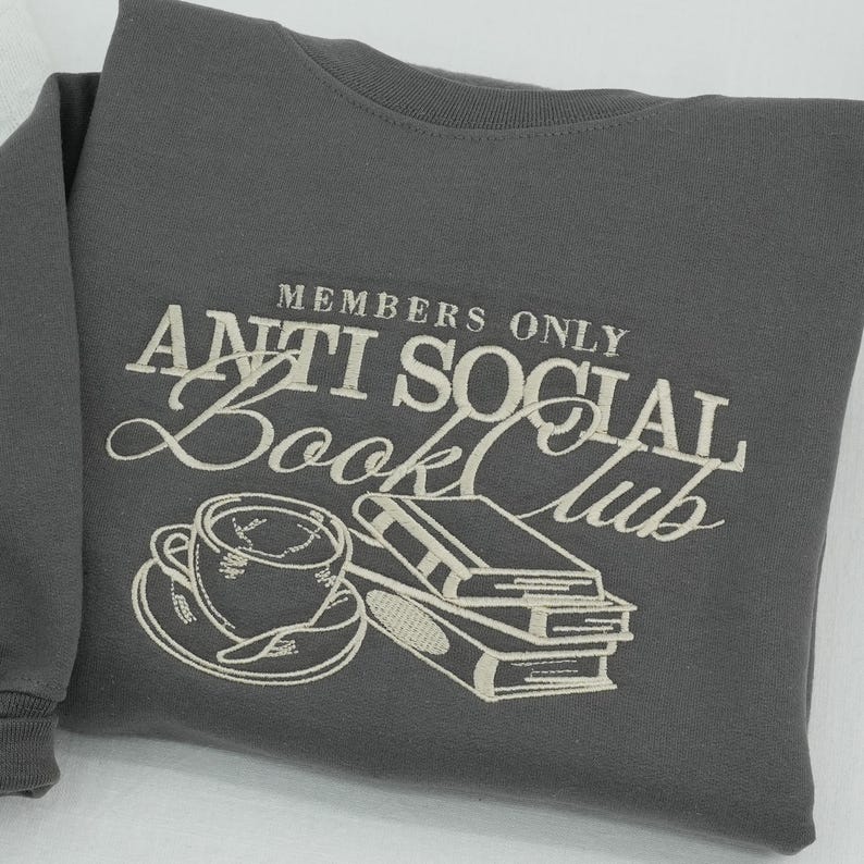 Macro detail of "Members Only Anti Social Book Club" embroidery on a charcoal grey Gildan sweatshirt. High-quality cream thread stitching showcases a realistic 3D relief effect of a coffee cup and stacked books. Professional commercial photography highlights the dense thread texture and premium craftsmanship. Ideal for showcasing custom embroidery quality on Etsy.