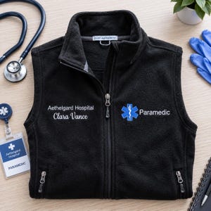 May include: A black fleece vest with a full-length front zip and two zipped pockets. The chest features white embroidery reading Aethelgard Hospital and Clara Vance, alongside a blue Star of Life emblem and the word Paramedic.