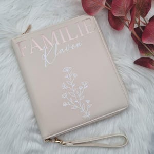 Könnte beinhalten: Eine beige Reißverschluss-Clutch mit dem Wort "FAMILIE" in Rosa und "Klavon" in weißer Schrift. Ein weißes Blumendesign befindet sich unten rechts. Die Clutch hat einen goldenen Reißverschluss und eine Handschlaufe.