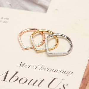 Op de afbeelding: Drie oorringen in roségoud, goud en zilver, elk met een witte opaal inleg in een V-vorm. De oorbellen worden getoond op een crèmekleurig oppervlak met de tekst "Merci beaucoup About Us".