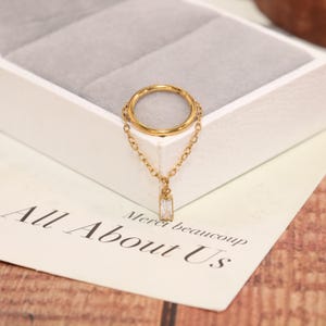 May include: A gold-toned hoop ring featuring a delicate attached chain that connects to a small, rectangular, clear baguette-cut stone charm. The jewelry piece is constructed with a circular band and a dangling pendant element.