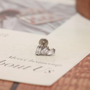 May include: A silver-toned flat back stud earring featuring three clear baguette-cut stones arranged in a stepped, staggered formation. The metal post is visible behind the decorative front, which is set with rectangular, faceted white crystals.