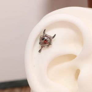 May include: A silver-toned metal ear piercing stud featuring a textured, irregular circular shape with two small horn-like protrusions at the top and a single marquise-cut red gemstone set in the center, resembling a stylized eye or creature.