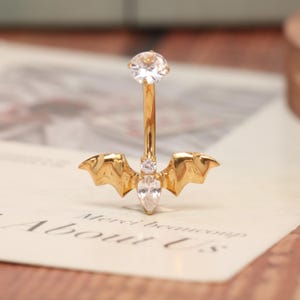 May include: Gold-colored belly button ring with a bat wing design. The ring features three clear, sparkling gemstones. The bat wings are detailed and add a unique touch to the jewelry.