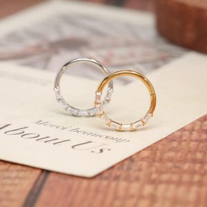May include: Two circular hinged rings, one in silver-tone metal and one in gold-tone metal, each featuring a row of clear baguette-cut stones set along the outer edge. The rings are positioned upright, showcasing their metallic finish and stones.