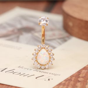 May include: A gold-coloured belly button ring. It features a clear, round gem at the top and a pear-shaped opal at the bottom, encircled by small, clear gems. The opal has a milky white appearance.